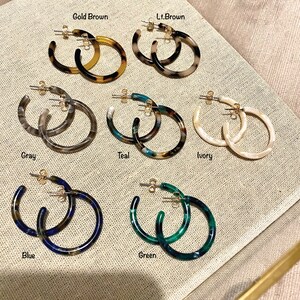 May include: Assortment of colorful hoop earrings made of tortoiseshell-like plastic. The earrings are displayed on a beige fabric surface. The colors include gray, teal, ivory, blue, green, light brown, and gold brown.
