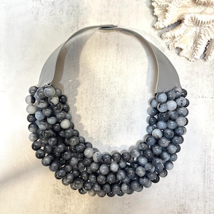 Puede incluir: Un collar de cuentas grises con una banda ancha de color gris similar al cuero. El collar presenta múltiples hilos de cuentas grises y blancas. Las cuentas son redondas y de diferentes tamaños. El collar se exhibe sobre una superficie clara.