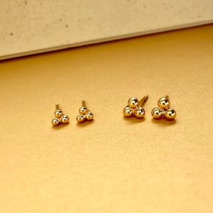 May include: Three pairs of gold-toned earrings with three small spheres clustered together in a triangular shape. The earrings are shown on a light brown surface.