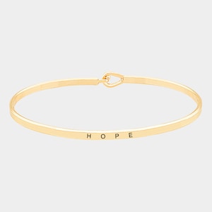 Puede incluir: Un brazalete dorado con la palabra "HOPE" grabada en la parte delantera.