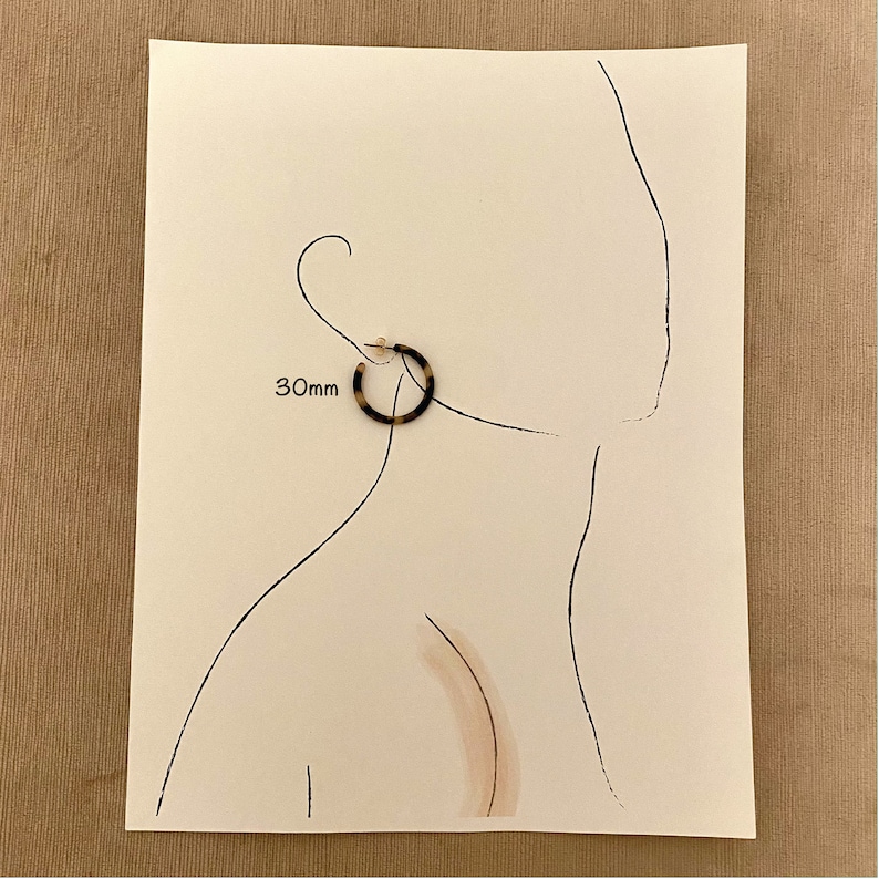 Op de afbeelding: Een paar bruine en zwarte creolen met een diameter van 30 mm. De oorbellen worden getoond op een witte achtergrond met een tekening van een oor.