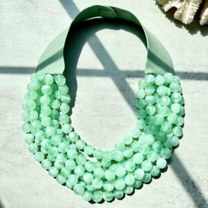 May include: A mint green statement necklace featuring multiple strands of faceted beads. The necklace has a wide, soft green band that sits around the neck. The beads are a light, translucent green color.