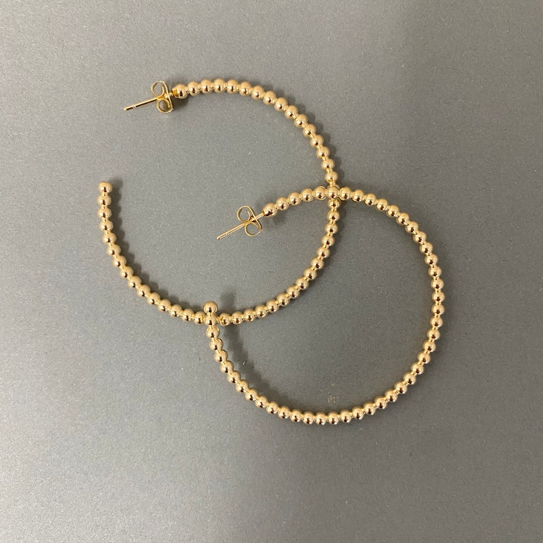 May include: A pair of gold hoop earrings made of small, round beads. The earrings are large and have a simple, elegant design.