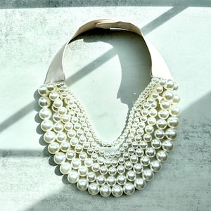 Può includere: Una collana a più fili con perle finte di diverse dimensioni. La collana ha un ampio colletto in nastro di raso chiaro. Le perle sono di un bianco crema, creando un look classico ed elegante.