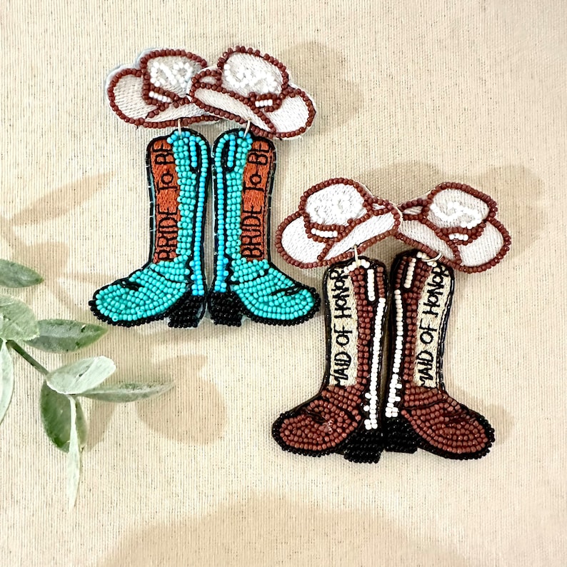 K&ouml;nnte beinhalten: Ein Paar Perlen-Cowboy-Stiefel-Ohrringe mit einem wei&szlig;en Cowboyhut-Anh&auml;nger. Ein Paar sagt "Bride to Be" und das andere sagt "Maid of Honor".