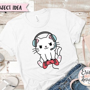 Op de afbeelding: Wit T-shirt met een cartoonillustratie van een witte kat die een blauwe en roze headset draagt en een rode videogamecontroller vasthoudt.