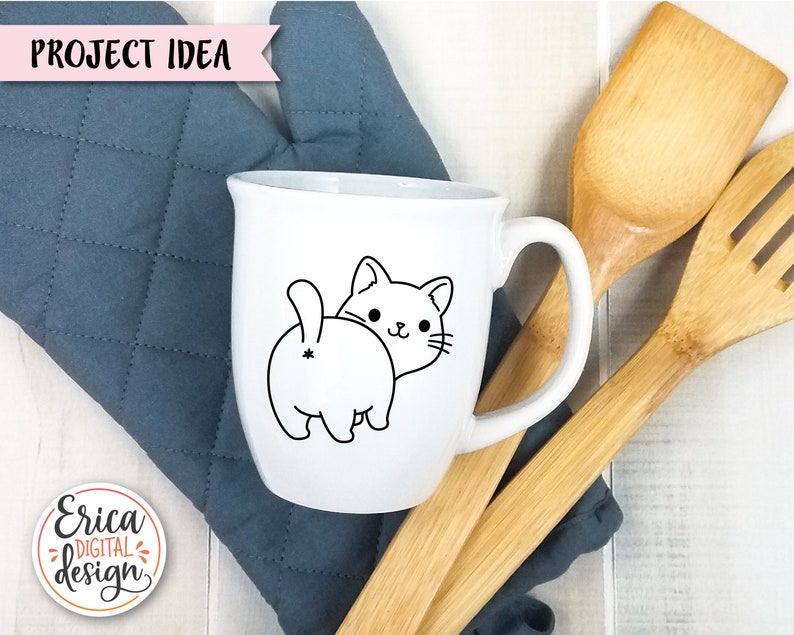 Puede incluir: Taza de cer&aacute;mica blanca con un dibujo de l&iacute;nea negra de la parte trasera y la cabeza de un gato. El gato mira hacia la derecha. La taza est&aacute; sobre un posavasos acolchado azul con utensilios de cocina de madera.