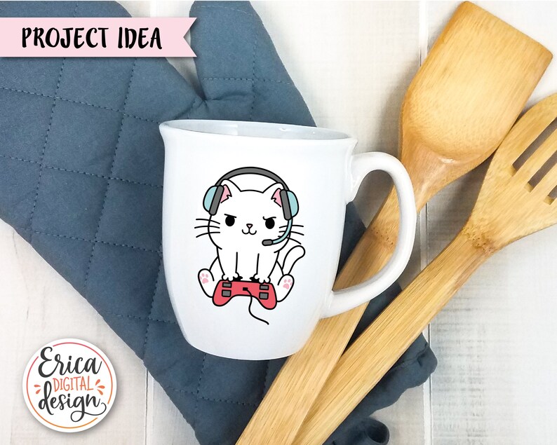 Op de afbeelding: Een witte keramische mok met een cartoonillustratie van een witte kat met een koptelefoon op en een rode videogamecontroller in zijn pootjes. De kat heeft blauwe ogen en een roze neus. De mok heeft een zwart handvat.