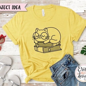Puede incluir: Una camiseta amarilla con un dibujo de l&iacute;nea negra de un gato con gafas durmiendo sobre una pila de libros. El texto "Project Idea" est&aacute; en una pancarta rosa en la parte superior de la imagen.
