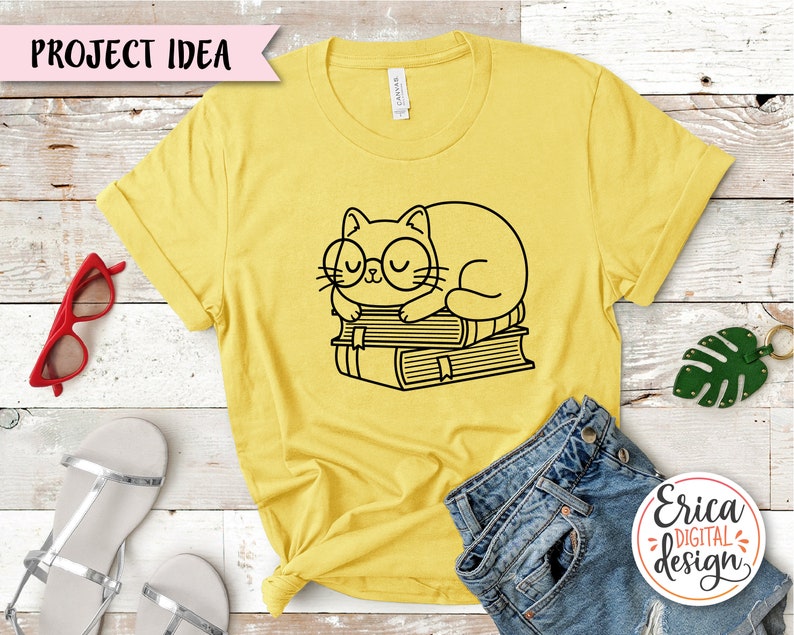 Puede incluir: Una camiseta amarilla con un dibujo de l&iacute;nea negra de un gato con gafas durmiendo sobre una pila de libros. El texto "Project Idea" est&aacute; en una pancarta rosa en la parte superior de la imagen.