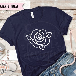 May include: A navy blue t-shirt with a white outline of a rose on the front. The shirt is laying on a wooden surface with a pair of jeans and a pink bag.
