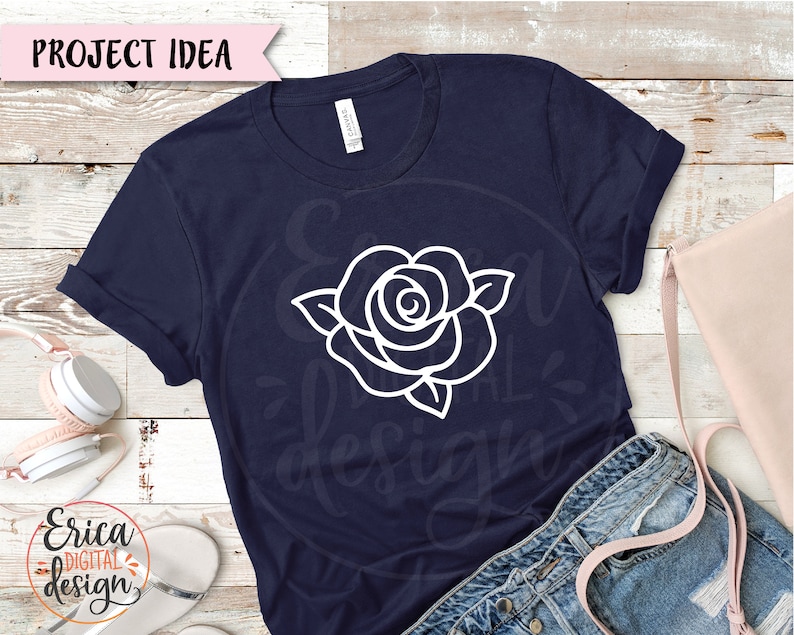 May include: A navy blue t-shirt with a white outline of a rose on the front. The shirt is laying on a wooden surface with a pair of jeans and a pink bag.