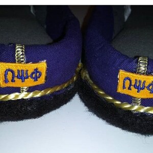 May include: A pair of purple slippers with black trim and gold rope detailing. Each slipper has a yellow patch with the letters "&Omega;&Psi;&Phi;" in blue.