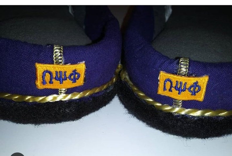 May include: A pair of purple slippers with black trim and gold rope detailing. Each slipper has a yellow patch with the letters "&Omega;&Psi;&Phi;" in blue.