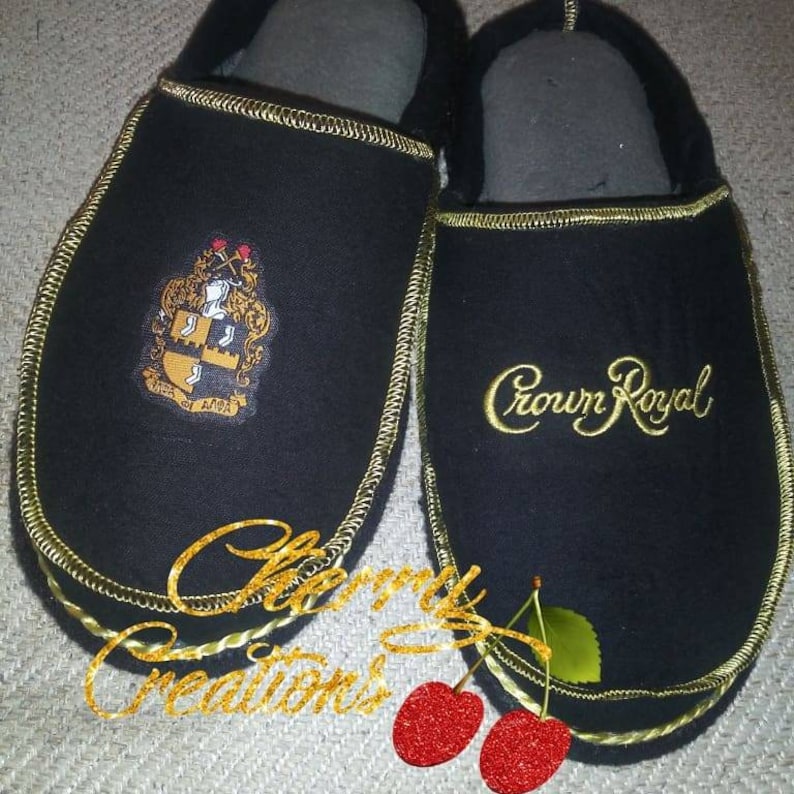May include: A pair of black slippers with gold trim. One slipper has a Crown Royal logo embroidered on it, and the other has a crest with the words "Alpha of Alpha".
