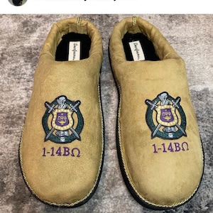 May include: A pair of tan suede slippers with black trim and a gold embroidered crest featuring a shield, swords, and the letters "N&Phi;&Phi;". The slippers also have the text "1-14&Beta;&Omega;" embroidered on the front.