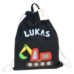 Pode incluir: Uma bolsa de cordão azul marinho com um cordão branco. A bolsa tem uma aplicação de veículo de construção vermelho e amarelo com o nome "LUKAS" em letras azuis.