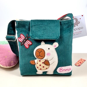 Puede incluir: Bolso bandolera de pana color turquesa con cierre de solapa y correa ajustable. Presenta un oso de dibujos animados aplicado con una barriga de lunares y una mariposa a cuadros rojos y blancos. El nombre "Emma" está bordado en el bolso.