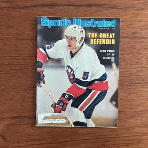 May include: A vintage Sports Illustrated magazine cover featuring a hockey player in a white, blue, and red uniform. The cover reads "Sports Illustrated" and "The Great Defender" with the player's name and team. The magazine is on a wooden surface.