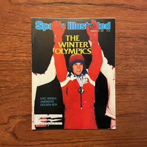 May include: Sports Illustrated magazine cover featuring Eric Heiden, America's Golden Boy, celebrating a Winter Olympics victory. The cover has a black background with the title "The Winter Olympics" in yellow and white. Heiden is wearing a red and white uniform.