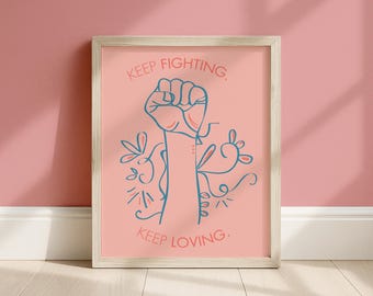 May include: A framed wall art print featuring a pink background with a central blue line drawing of a raised fist. The text "KEEP FIGHTING." appears above the fist, and "KEEP LOVING." is printed below it, surrounded by blue floral line art.