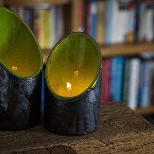 Könnte beinhalten: Zwei schwarze und grüne Kerzenhalter aus Keramik mit einer gelben Kerzenflamme in jedem. Die Kerzenhalter stehen auf einer Holzoberfläche.