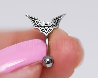 May include: A silver belly button ring with a detailed bat charm. The ring is held by a finger with pink nail polish.