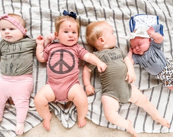 May include: Four babies wearing different graphic tees lay on a striped blanket. The tees say "Adventure is out there", "Peace", "Tree Hugger", and "Wild".