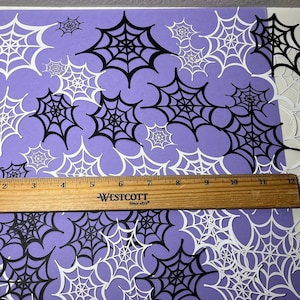 May include: A sheet of purple paper with black and white paper cutouts of spiderwebs. The paper cutouts are of various sizes and are arranged in a random pattern. A ruler is placed at the bottom of the sheet to show the size of the cutouts.
