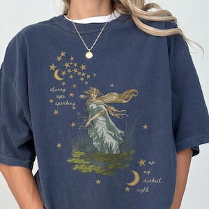 May include: Navy blue t-shirt featuring a vintage illustration of a woman with long hair, stars, and a crescent moon. The text on the shirt says "starry eyes sparkling" and "up my darkest night."