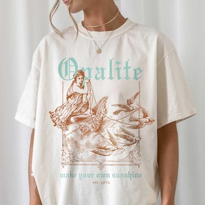 Pode incluir: T-shirt creme com um gráfico de estilo vintage. O design apresenta a palavra "Qualite" em verde-azulado, juntamente com uma ilustração de uma mulher e a frase "make your own sunshine" em verde-azulado. A camisa tem um corte folgado.
