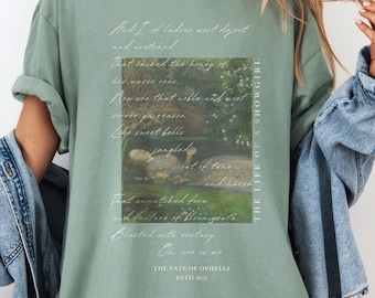 May include: Sage green t-shirt featuring a printed image of a painting and script. The text on the shirt includes the phrase "THE LIFE OF A SHOWGIRL" and "THE FATE OF OPHELIA ESTD 1621". The shirt has a relaxed fit.