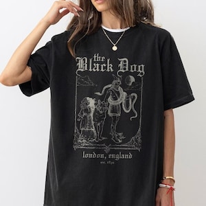 May include: Black t-shirt with a graphic print of a man with a snake, a dog, and a woman. The text "the Black Dog" is printed above the graphic. The text "london, england est. 1820" is printed below the graphic.