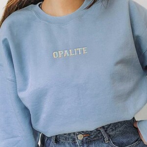 May include: A light blue sweatshirt with the word "OPALITE" embroidered in a lighter color across the chest. The sweatshirt is paired with blue denim jeans. The model is wearing the sweatshirt and jeans.