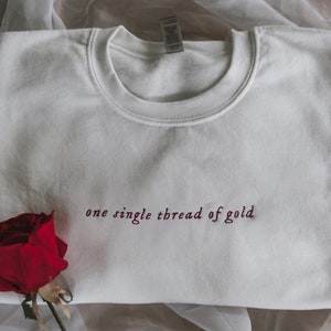 May include: White sweatshirt with the text "one single thread of gold" embroidered in maroon on the front. A red rose is in the bottom left corner of the image.