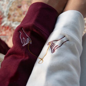 May include: Two sleeves of a sweatshirt, one burgundy and one white, with embroidered hands reaching towards each other, connected by a gold thread.