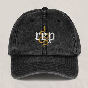 Peut inclure: Casquette de baseball noire de style vintage avec le mot "rep" brodé en lettres gothiques blanches. Un serpent doré est brodé verticalement à travers les lettres. La casquette a une visière incurvée et un bouton noir sur le dessus.