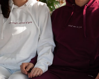 Puede incluir: Dos sudaderas con capucha a juego, una blanca con el texto "not a single thread of gold" en burdeos, y la otra burdeos con el texto "tied me to you" en blanco.