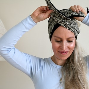 Könnte beinhalten: Eine Person in einem hellblauen Langarmshirt bindet sich einen schwarzen und braunen Leopardenmuster-Haar-Turban um den Kopf.