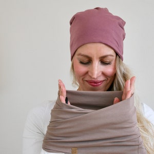 Könnte beinhalten: Eine altrosa Beanie-Mütze und ein dazu passender taupefarbener Infinity-Schal. Die Mütze besteht aus einem einfarbigen, glatten Stoff mit abgerundeter Form. Der Infinity-Schal ist ein breites, schlauchförmiges Stoffstück, das in Lagen um den Hals liegt.