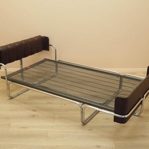 Op de afbeelding: Een vintage slaapbank met een donkerbruin lederen hoofdsteun en armleuningen. Het bedframe is gemaakt van verchroomd metaal en de matrasdrager is een zwart metalen gaas. De slaapbank staat tegen een neutraal gekleurde muur.