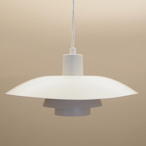 Könnte beinhalten: Eine weiße Pendelleuchte mit schlichtem, modernem Design. Die Leuchte hat einen runden, flachen Schirm mit drei Stufen und einem weißen Kabel.