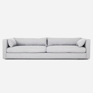 Op de afbeelding: Een lichtgrijze, moderne bank met twee grote zitkussens en twee rugkussens. De bank heeft een laag profiel en twee kleinere sierkussens aan elk uiteinde. De stof lijkt een geweven textuur te hebben.
