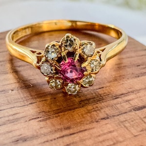 May include: A gold ring with a central pink gemstone encircled by smaller, clear stones. The ring's band is polished gold, and the setting has an ornate, floral design. The ring is displayed on a wooden surface.