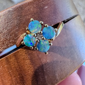 Op de afbeelding: Een gouden ring met vier ronde, iriserende blauwe opalen in een clusteropstelling, met een kleine, heldere, rondgeslepen steen in het midden. De band is gemaakt van gepolijst geelgoud en heeft een glad, gebogen ontwerp dat naar de zetting toe loopt.
