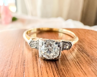 Pode incluir: Um anel de ouro com um diamante central de corte redondo engastado numa cabeça quadrada de metal branco. Os ombros do anel são acentuados com diamantes mais pequenos em trabalhos metálicos decorativos, criando um aspeto detalhado.