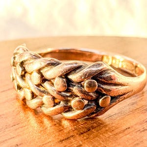 May include: A gold-colored ring with a braided design. The ring features a series of interwoven strands, creating a textured surface. Small, round accents are evenly spaced along the braid. The ring is set against a blurred background.