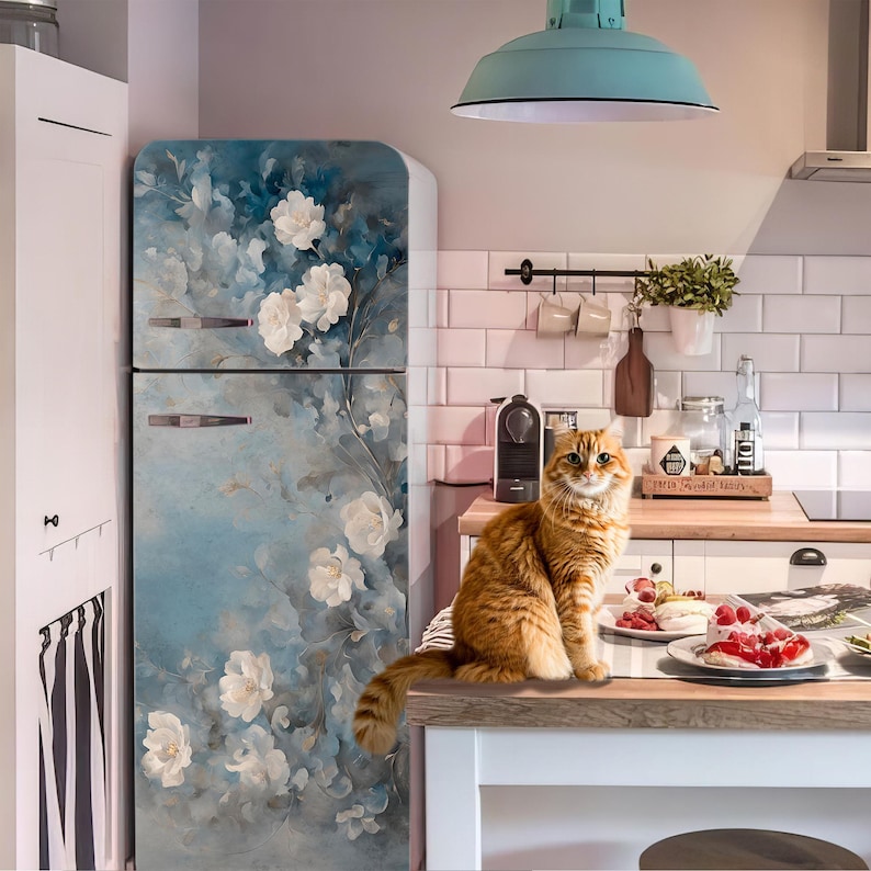 May include: A retro-style refrigerator with a floral design in shades of blue and white. An orange tabby cat sits on the countertop in front of the refrigerator. The kitchen has a vintage aesthetic with a light blue pendant lamp.