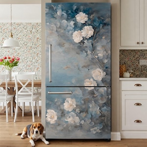 Op de afbeelding: Een koelkast met een bloemenontwerp. De voorkant van het apparaat is beschilderd met een aquarel-achtig beeld van witte bloemen en bladeren tegen een blauw-grijze achtergrond. De koelkast heeft een zilveren handvat.