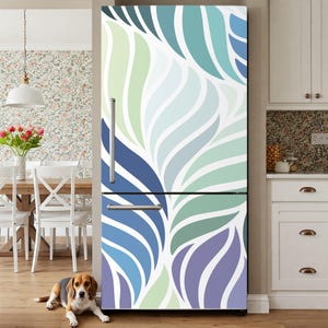 Op de afbeelding: Een koelkast met een kleurrijk bladpatroon in tinten blauw, groen en paars. Het ontwerp bedekt de hele voorkant van het apparaat. De koelkast staat in een keuken met een hond die op de grond ligt.
